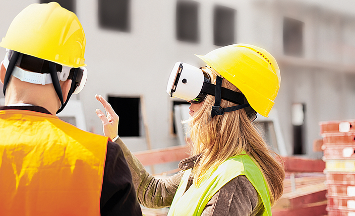 Eine Frau mit Schutzhelm und Vier-Punkt-Kinnriemen trägt eine virtuelle Brille (VR-Brille) auf einer Baustelle.
    