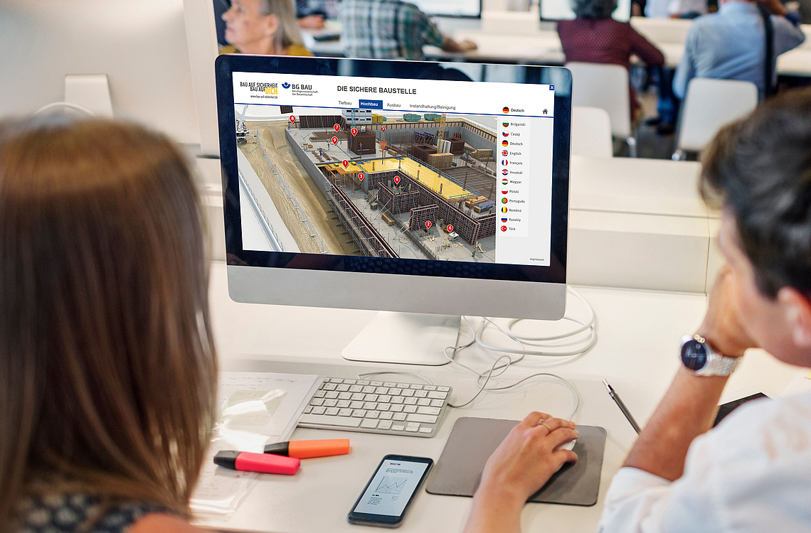 Zwei Personen schauen auf einen Monitor. Es ist darauf eine Baustelle zu sehen.
    