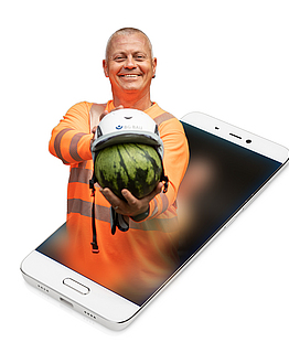 Die Aufsichtsperson Denny Hillert zeigt anhand einer Melone wie gut ein Schutzhelm den Kopf schützt. Sein Oberkörper ist auf ein Smartphone montiert.
    