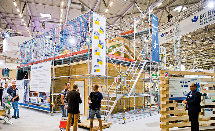 Messestand mit offenem Holzhaus, an dem draußen Gerüste und Treppen angebracht sind.
    