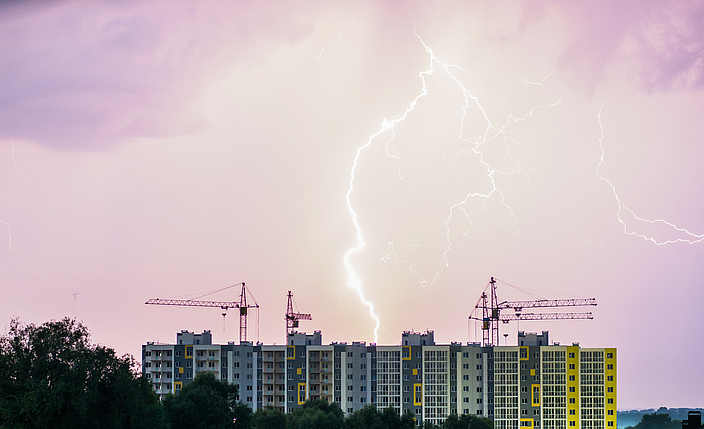 Ein Mehrfamilienhaus mit Baukränen vor dem Hintergrund des Nachthimmels, wo man Blitze am Himmel sehen kann.
    